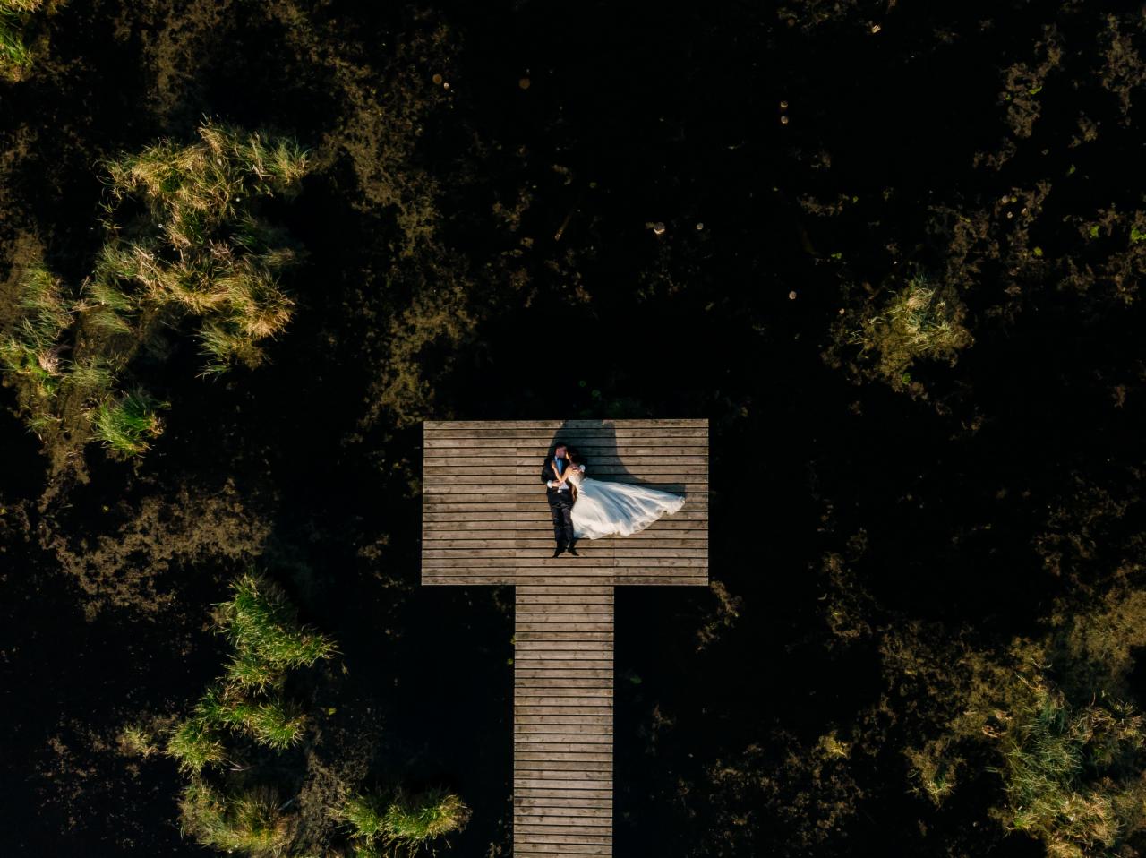 Para młoda leżąca na drewnianym pomoście w otoczeniu ciemnej wody i roślinności, widok z lotu ptaka.