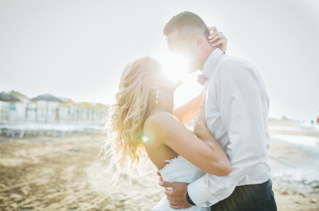 Para młoda w objęciach na plaży, silne światło słoneczne, panna młoda z blond lokami i długimi kolczykami, w tle budynki plażowe.