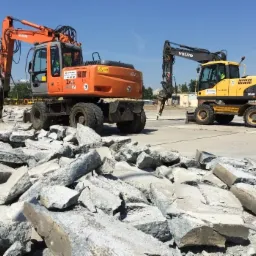 Dwie koparki kołowe, Hitachi Zaxis 160W i Volvo EW160C, z młotami hydraulicznymi na placu budowy, gruz betonowy w pierwszym planie, w tle budynek i żuraw budowlany.