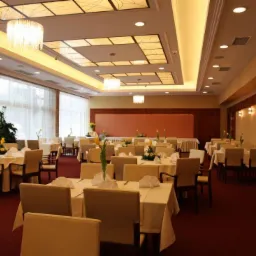 Uroczysta sala bankietowa z nakrytymi stołami, dekoracjami kwiatowymi i eleganckim oświetleniem sufitowym. Przy oknach widoczne firany, a na stołach białe obrusy i serwetki.