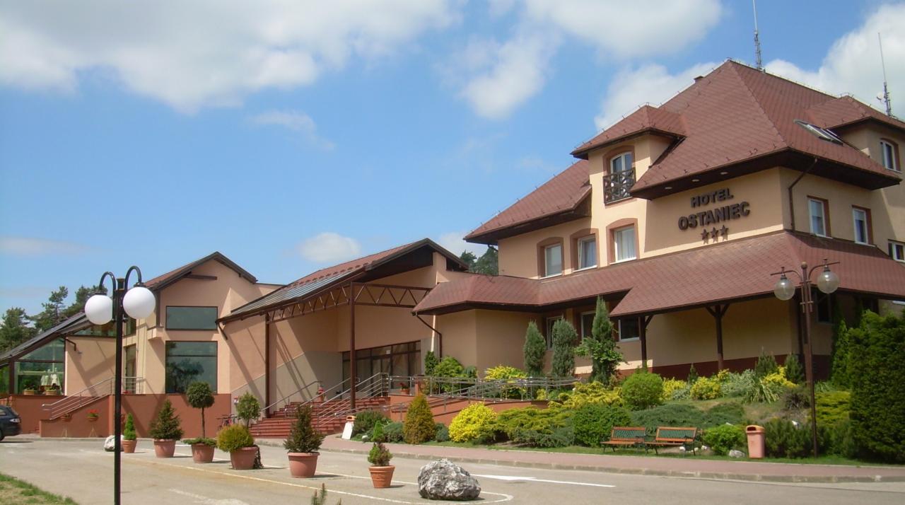 Fasada hotelu z brązowym dachem i jasną elewacją, widoczny napis 'Hotel Ostaniec ***', zadbany ogród z krzewami i ławeczkami przed budynkiem, lampy uliczne z kulistymi kloszami.