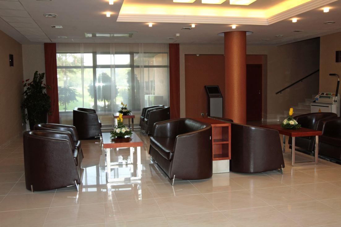 Lobby hotelu SPA z skórzanymi fotelami, stolikami z kwiatami i lśniącą podłogą z płytek, widok na jasne okna i kolumnę.