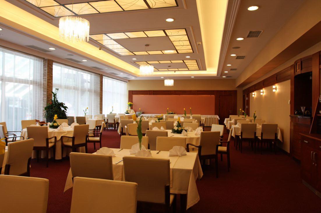 Uroczysta sala bankietowa z nakrytymi stołami, dekoracjami kwiatowymi i eleganckim oświetleniem sufitowym. Przy oknach widoczne firany, a na stołach białe obrusy i serwetki.