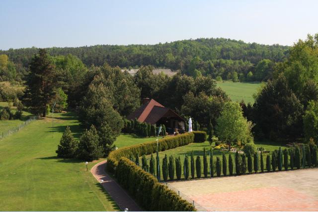 Widok z góry na zadbany teren zielony z budynkiem z czerwonym dachem, otoczonym żywopłotem i drzewami, w tle las.