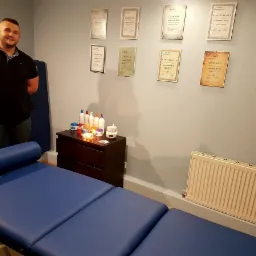 Gabinet z niebieskim łóżkiem do zabiegów, komodą z kosmetykami i certyfikatami na ścianie, na tle których stoi uśmiechnięty mężczyzna.