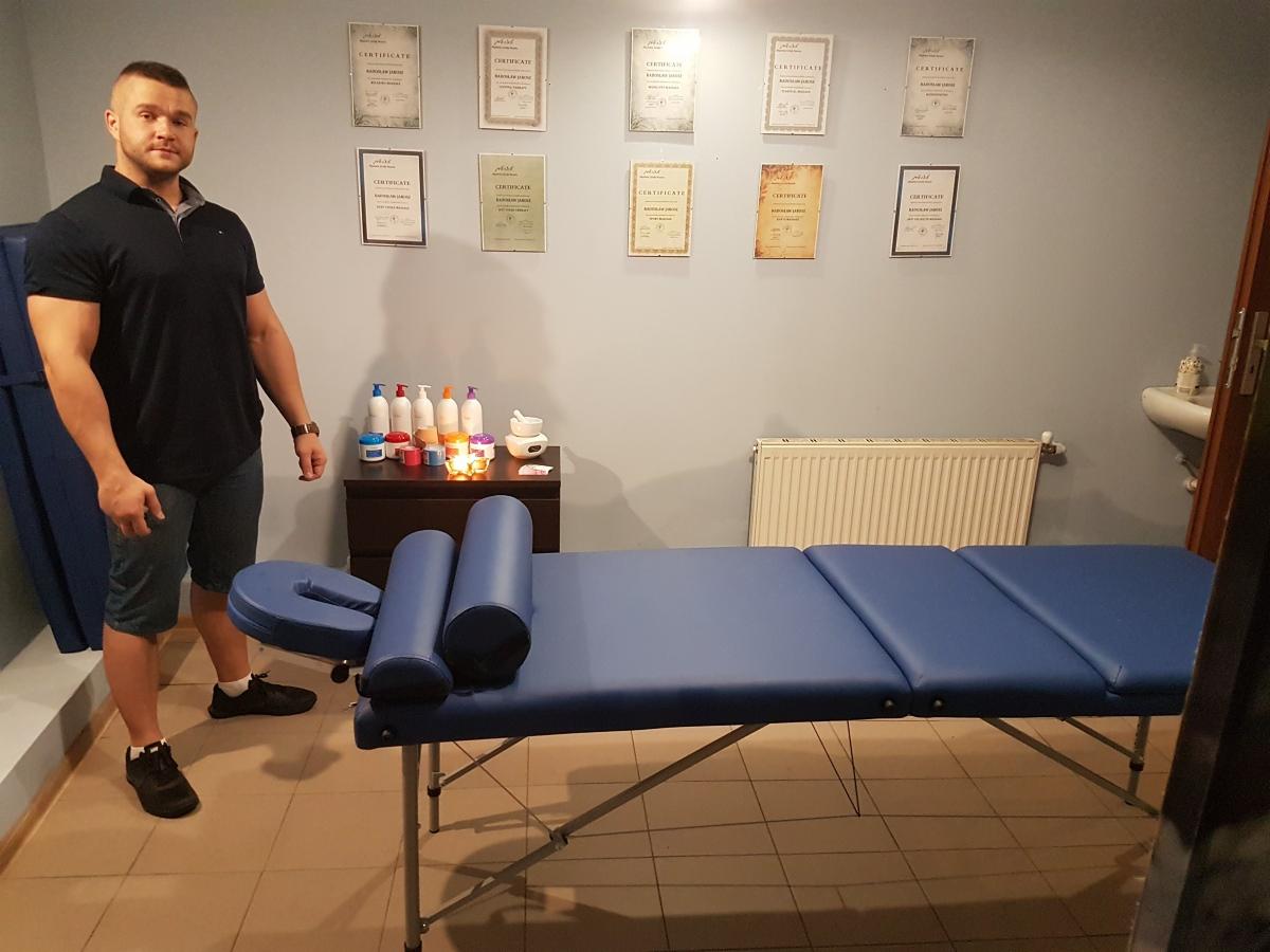 Mężczyzna stoi obok niebieskiego stołu do zabiegów, na ścianie wiszą certyfikaty, na szafce kosmetyki i świeczki.