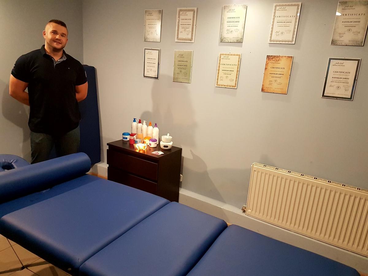 Gabinet z niebieskim łóżkiem do zabiegów, komodą z kosmetykami i certyfikatami na ścianie, na tle których stoi uśmiechnięty mężczyzna.