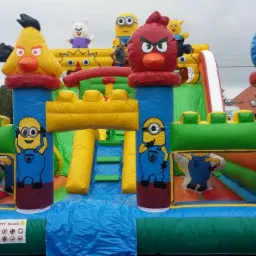 Duży, kolorowy dmuchany zamek ze zjeżdżalnią, ozdobiony postaciami z bajek i gier, ustawiony na trawniku przed domem.