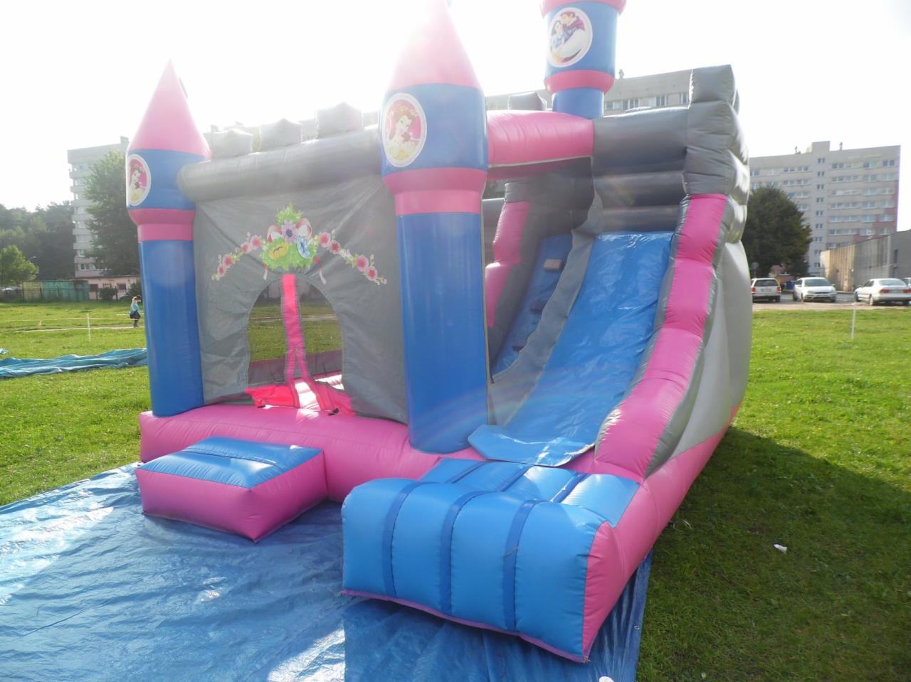 Rozłożony, dmuchany zamek z motywem księżniczki, w odcieniach różu, błękitu i szarości, ustawiony na trawie i niebieskiej plandece.