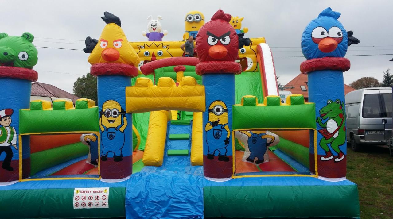 Duży, kolorowy dmuchany zamek ze zjeżdżalnią, ozdobiony postaciami z bajek i gier, ustawiony na trawniku przed domem.