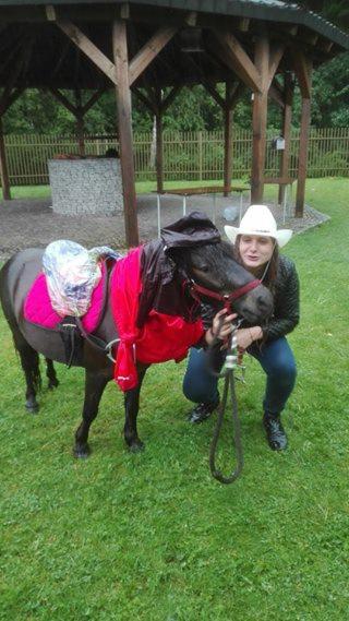 Kobieta w kowbojskim kapeluszu kuca obok małego konia ubranego w różowe siodło i pelerynę, na trawniku pod drewnianą altaną.