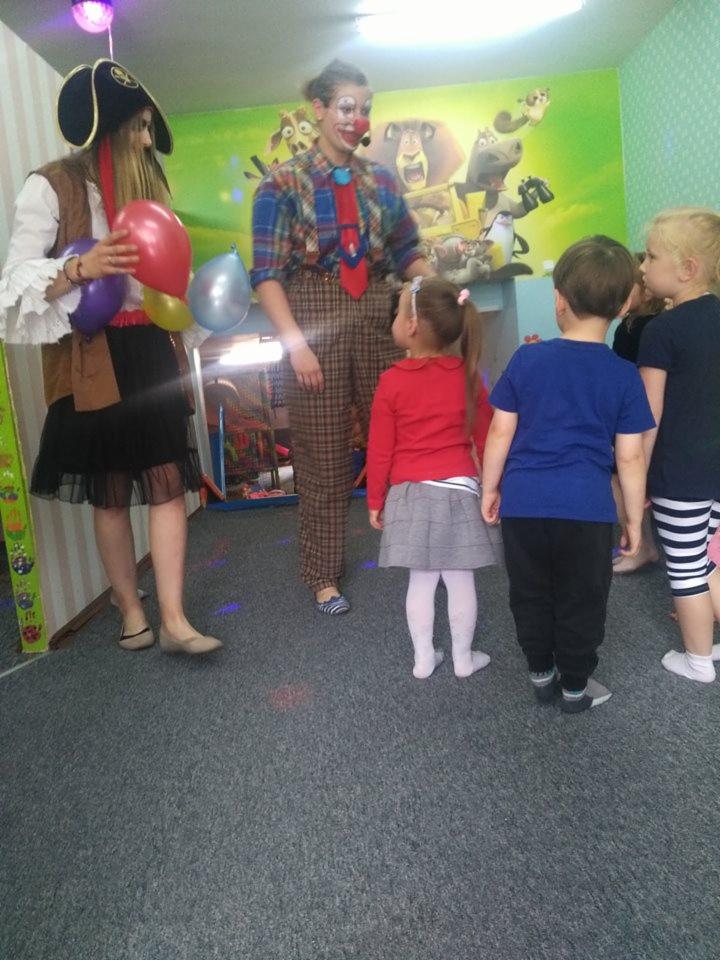 Dwie osoby przebrane za pirata i klauna, stoją przed grupą dzieci w pokoju z zieloną ścianą i postaciami z bajek