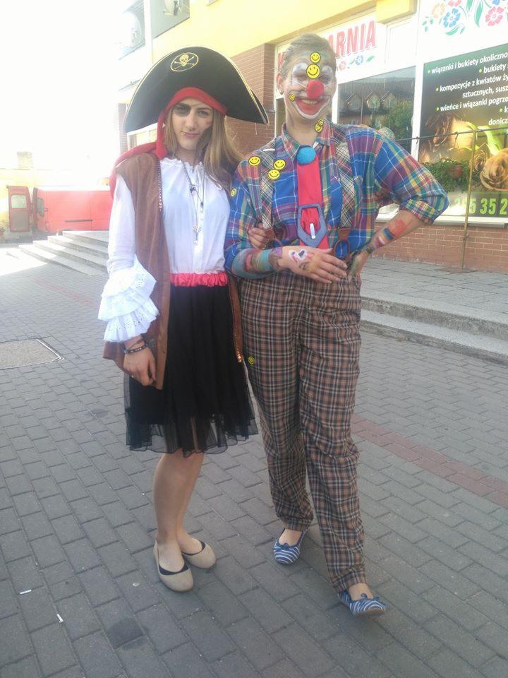 Dwie osoby przebrane: jedna za pirata z czarną opaską na oku i kapeluszem, druga za klauna z pomalowaną twarzą i w kraciastym ubraniu, stoją na chodniku przed budynkiem.