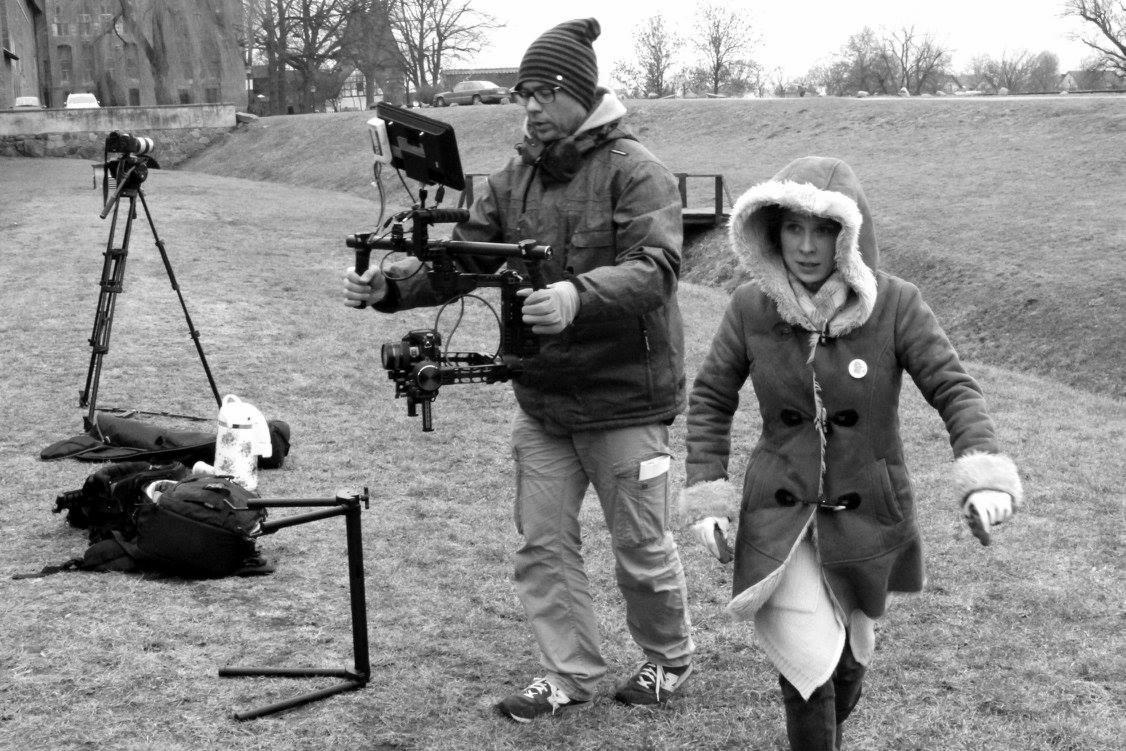 Czarno-białe zdjęcie z planu filmowego: kamerzysta z profesjonalnym sprzętem i aktorka w zimowym płaszczu na łące, w tle budynek i drzewa.