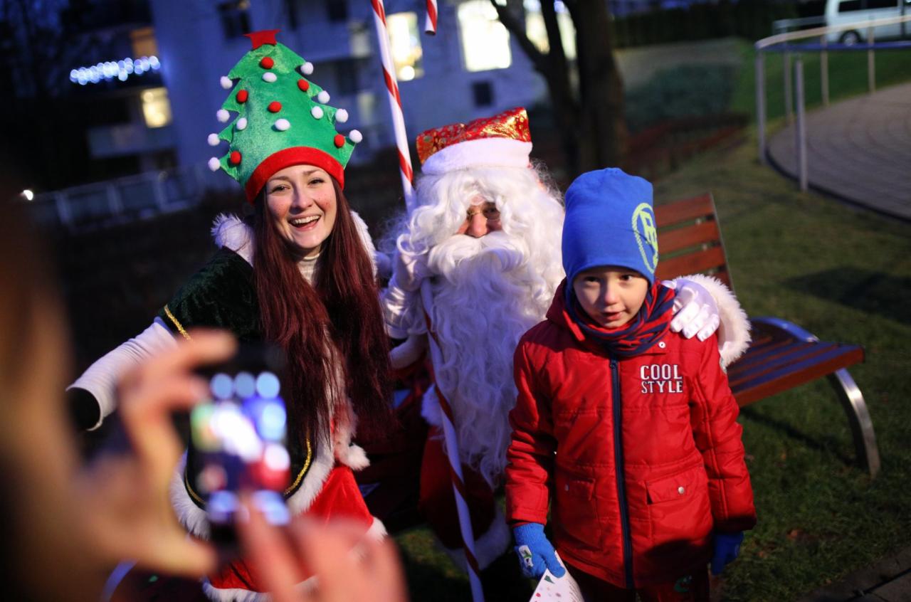 Uśmiechnięta kobieta w kostiumie choinki z lizakiem, Święty Mikołaj z brodą i chłopiec w czerwonej kurtce pozują do zdjęcia robionego smartfonem na tle parku.