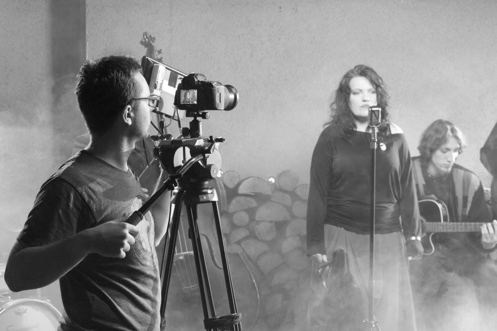 Czarno-białe zdjęcie z planu filmowego: kamerzysta z kamerą na statywie, wokalistka przed mikrofonem i gitarzysta w tle, w otoczeniu scenografii z drewnem i dymem.