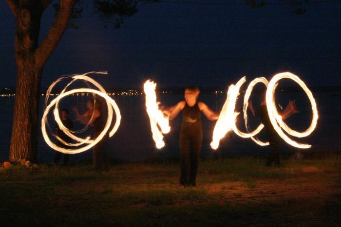 Pokaz fire show: artysta w ciemnym stroju wiruje płonącymi poi, tworząc świetlne okręgi na tle nocnego krajobrazu z drzewem i jeziorem.