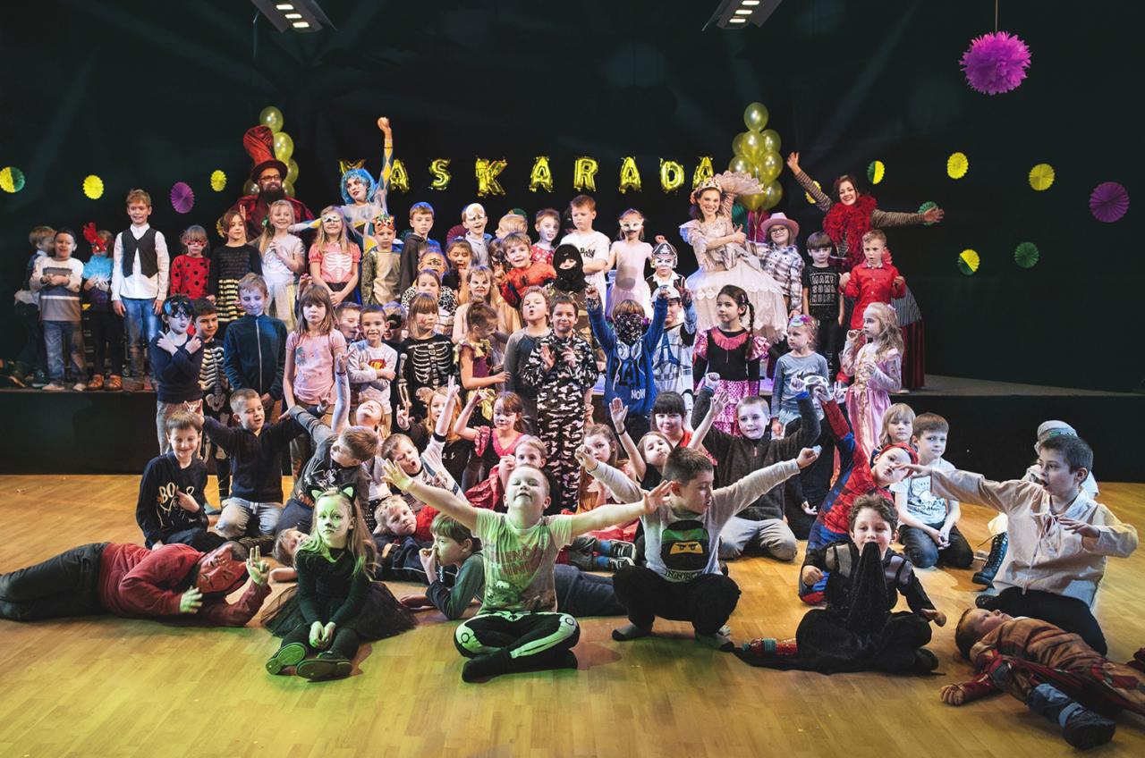 Grupa dzieci i dorosłych w kostiumach na scenie podczas przedstawienia maskarady, z dekoracjami z balonów i papierowych ozdób.