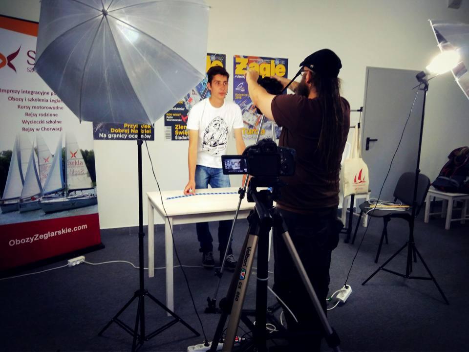 Kamerzysta filmuje młodego mężczyznę w studio, widoczny parasol oświetleniowy, kamera na statywie i plakaty żeglarskie w tle.