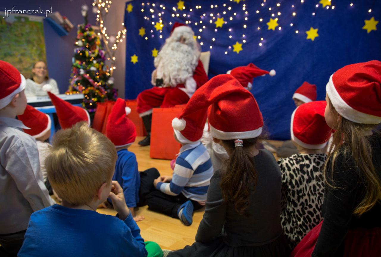 Dzieci w czerwonych czapkach Mikołaja siedzą na podłodze i patrzą na Mikołaja siedzącego na krześle przed niebieskim tłem z gwiazdami i choinką, w tle widoczna kobieta.