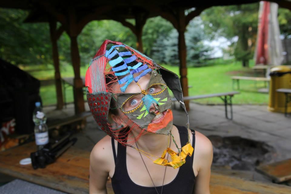 Osoba z kolorową, ręcznie robioną maską na twarzy, z widocznymi elementami tkaniny, papieru i farby, na tle altany i zieleni.