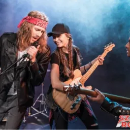 Zespół rockowy w skórzanych kurtkach gra na scenie z dymem w tle, widoczne gitary, mikrofon i perkusja, logo szkoły rocka w prawym dolnym rogu.