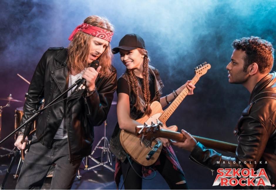 Zespół rockowy w skórzanych kurtkach gra na scenie z dymem w tle, widoczne gitary, mikrofon i perkusja, logo szkoły rocka w prawym dolnym rogu.
