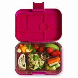 Otwarty, różowy lunchbox z przegródkami wypełniony różnorodnymi przekąskami: kostki sera z bazylią, pomidorki cherry, wrap z szynką, makaron farfalle z pesto, mieszanka nasion, truskawki, gruszka...