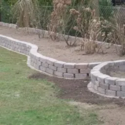 Kaskadowo ułożona, niska ścianka z kostki brukowej oddzielająca trawnik od rabaty z suchymi roślinami. Widoczne świeżo dosypana ziemia i fragment okrągłej rabaty.