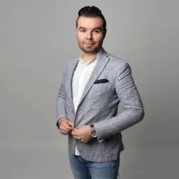 Mężczyzna w jasnej, kraciastej marynarce zapinający guzik, ubrany w białą koszulę i jeansy, z zegarkiem na ręku, na szarym tle.