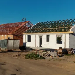 Dwa budynki w trakcie budowy: jeden z czerwonej cegły z drewnianą konstrukcją dachu, drugi z białych pustaków z częściowo wykonaną więźbą dachową. Na pierwszym budynku widać pracownika.