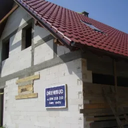 Niedokończony dom murowany z czerwonym dachem i oknem dachowym, widoczne pustaki, betonowe nadproża, prowizoryczne zabezpieczenia okien i drzwi deskami, szyld firmy budowlanej Drewbud.