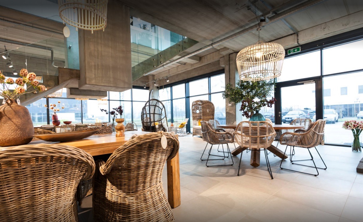 Wnętrze przestronnego salonu z betonowym stropem i dużymi oknami, ukazujące drewniany stół otoczony wiklinowymi krzesłami, wiszące lampy z naturalnych materiałów i dekoracje roślinne w wazonach.