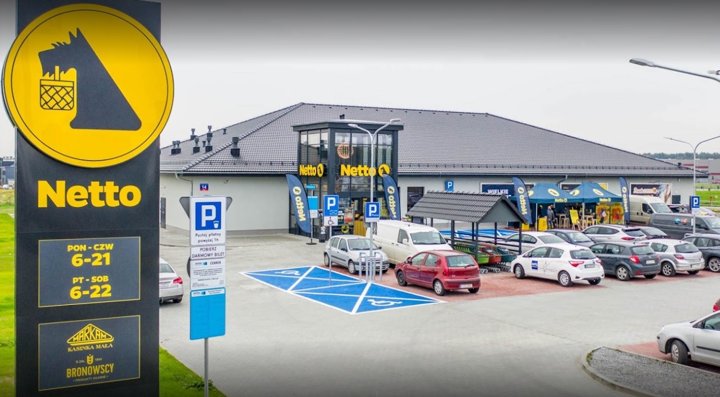 Widok zewnętrzny sklepu Netto w Zatorze z parkingiem, na którym widać kilka samochodów. Duży żółto-czarny szyld z logo sklepu i godzinami otwarcia.
