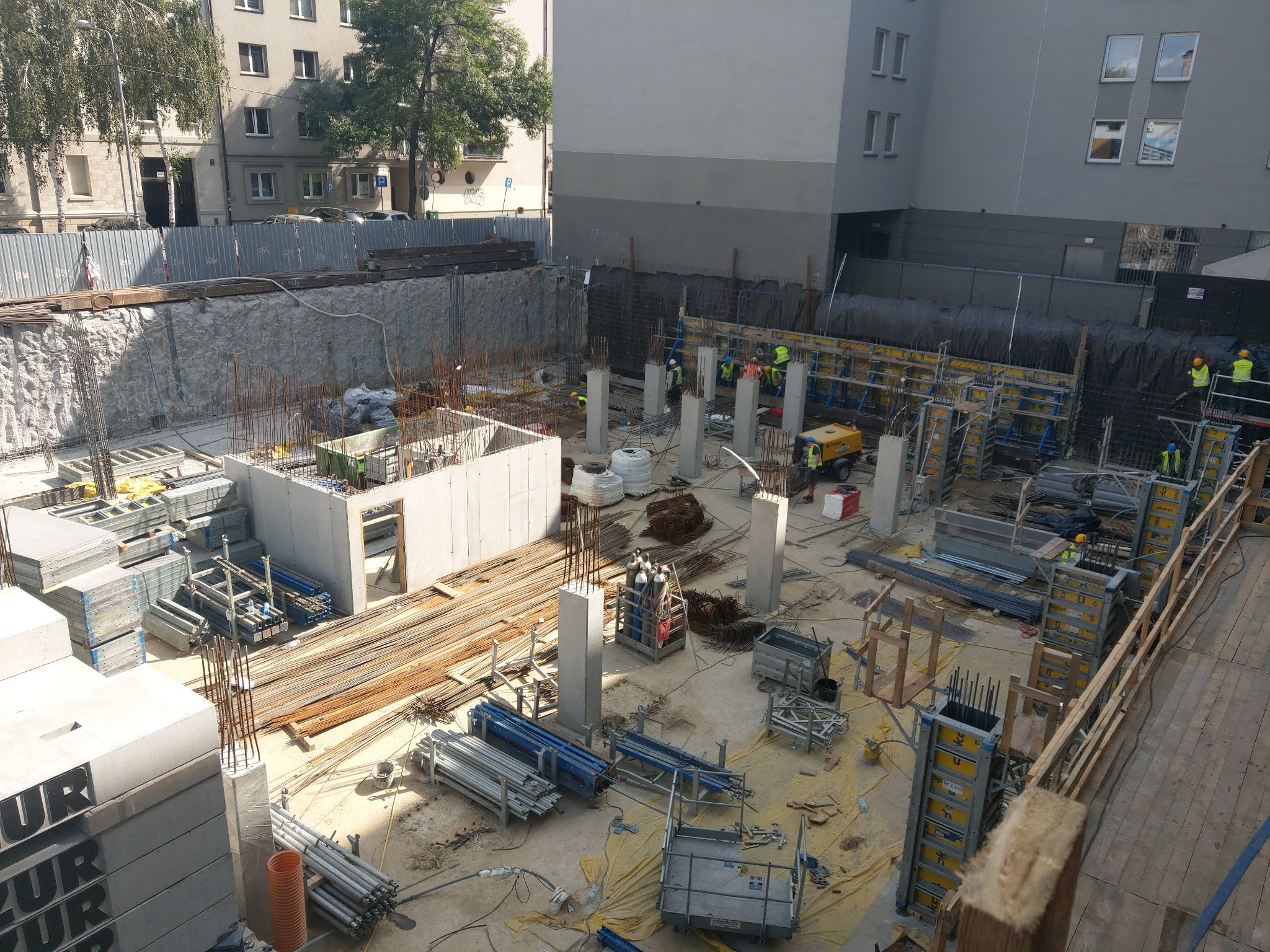 Widok z góry na plac budowy z fundamentami, szalunkami, zbrojeniami i pracownikami w kamizelkach odblaskowych, przygotowującymi teren pod budynek mieszkalny w miejskiej zabudowie.