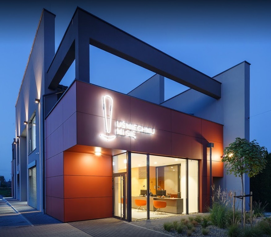 Nowoczesny budynek usługowy z elewacją z paneli w odcieniach szarości i terakoty, przeszklonym wejściem i neonowym logo firmy 'Uśmiechnij Mi Się' nad wejściem, sfotografowany o zmierzchu.