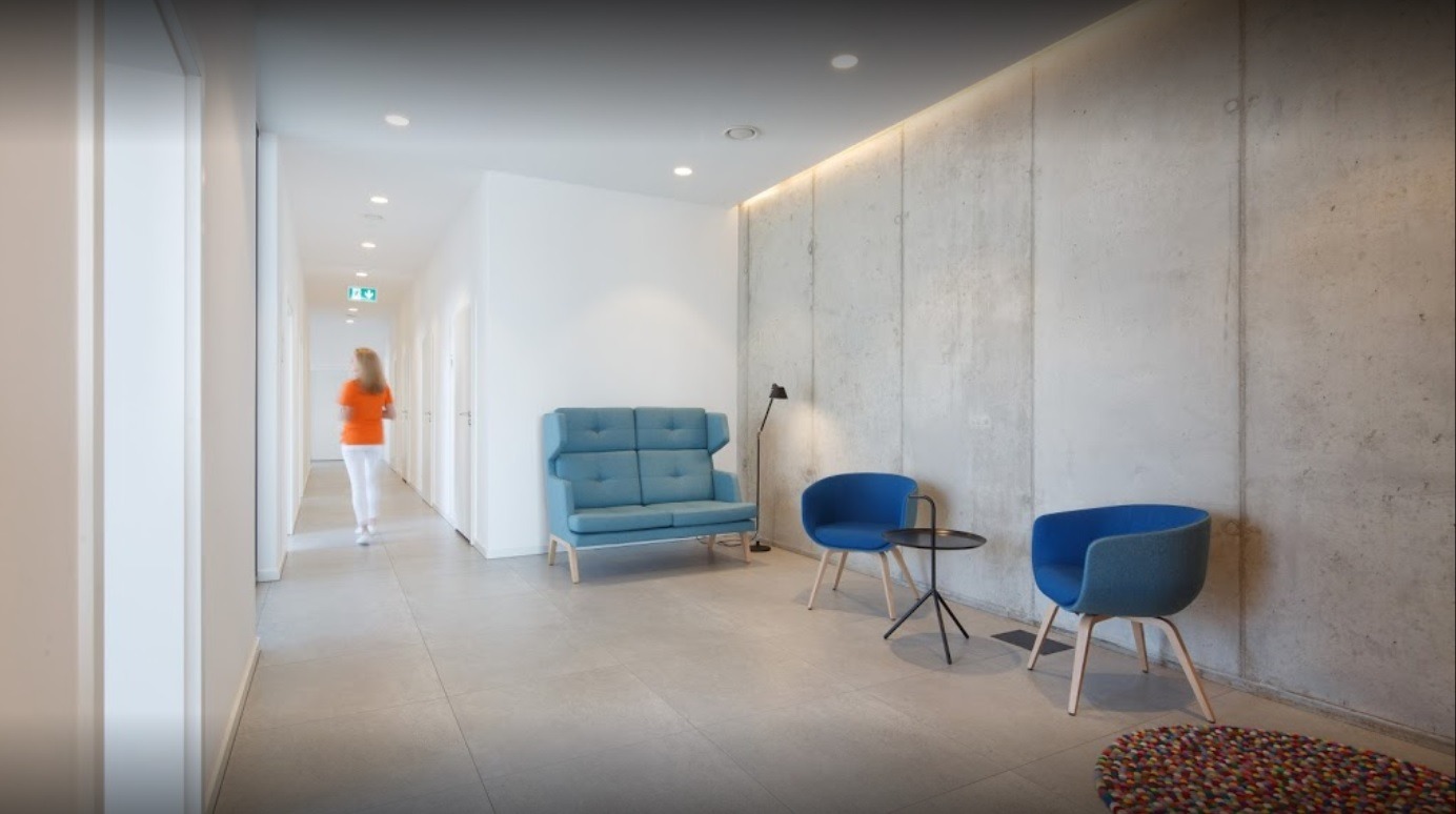 Nowoczesne wnętrze korytarza z betonową ścianą, błękitnymi meblami do siedzenia i przechodzącą osobą w pomarańczowej koszulce.