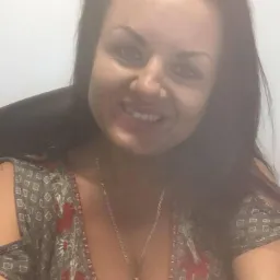 Portret uśmiechniętej kobiety z ciemnymi włosami w bluzce z wzorem kwiatowym, z łańcuszkiem na szyi, na tle jasnej ściany.