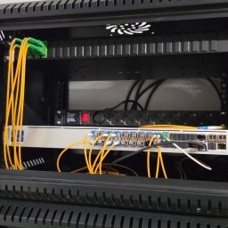Proste IT Sp. z o.o. - Otwarta szafa rack z patch panelem, switchami i okablowaniem światłowodowym. Żółte kable patch cord. Widoczny wentylator chłodzący. Instalacja sieci komputerowej.