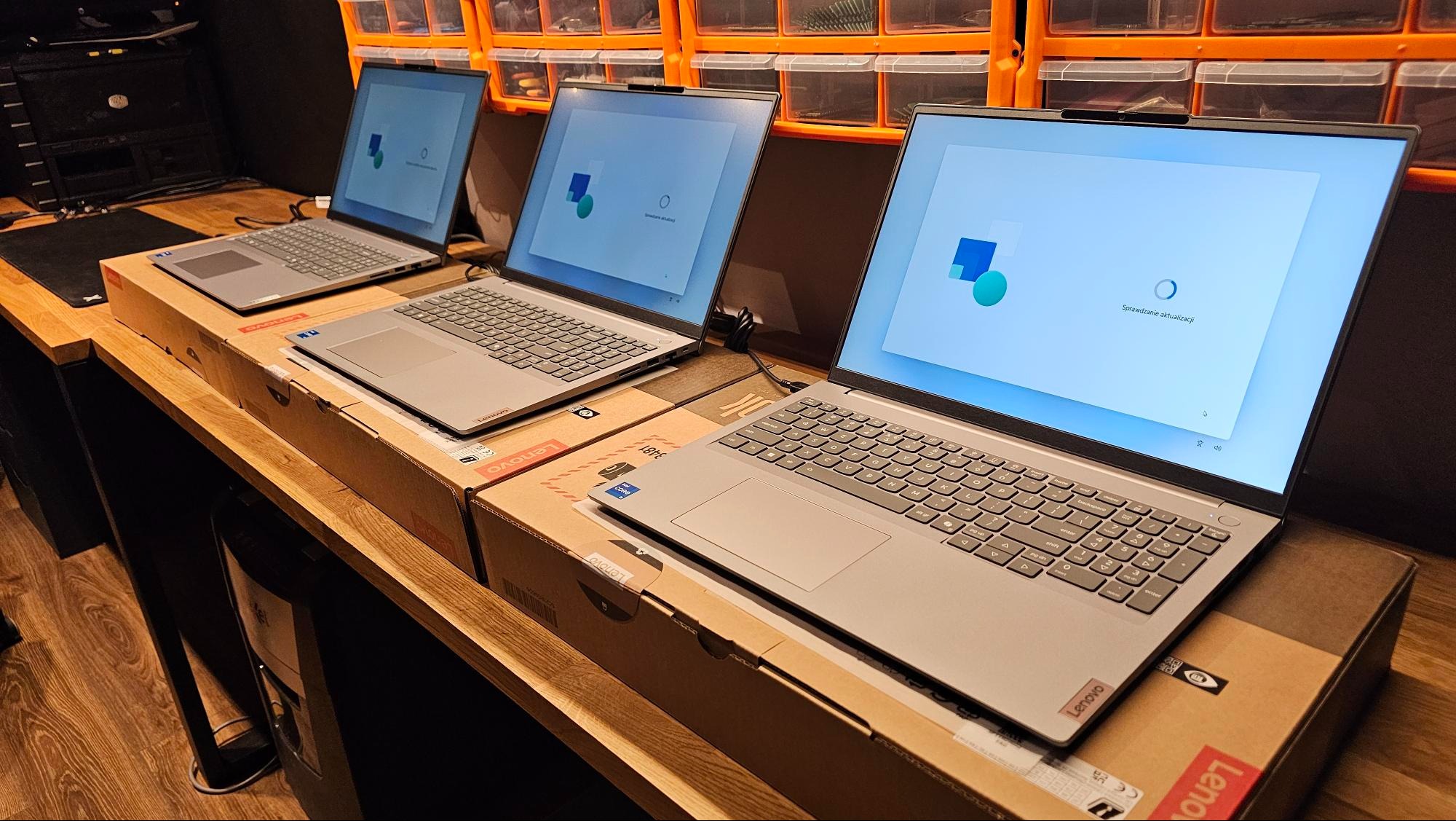 Trzy szare laptopy Lenovo na kartonach, z ekranami w trakcie aktualizacji systemu Windows, ustawione na drewnianym biurku w pomieszczeniu z pomarańczowymi pojemnikami.