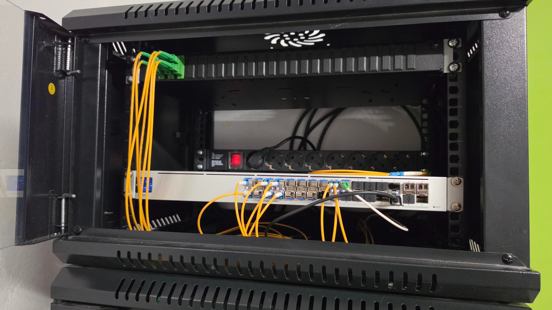 Otwarta szafa rack z patch panelem, switchami i okablowaniem światłowodowym. Żółte kable patch cord. Widoczny wentylator chłodzący. Instalacja sieci komputerowej.