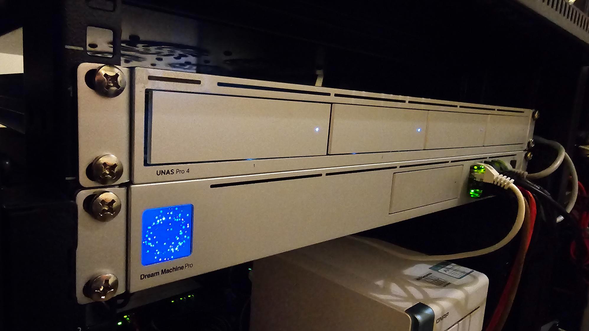 Serwer NAS UNAS Pro 4 i Dream Machine Pro w szafie rack, z podłączonymi kablami sieciowymi. Widoczne diody sygnalizujące pracę urządzeń.
