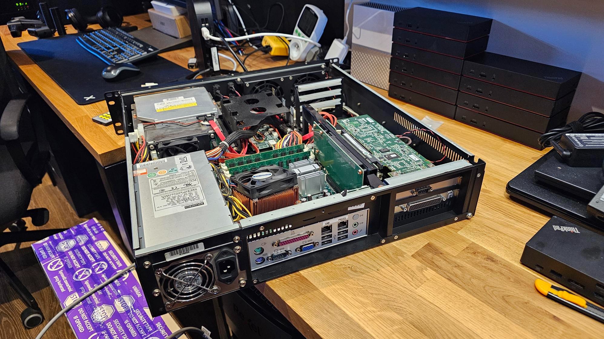 Otwarta obudowa serwera rack z widocznymi komponentami: płyta główna, karty rozszerzeń, zasilacz. Obok stos serwerów ThinkPad i narzędzia na biurku.