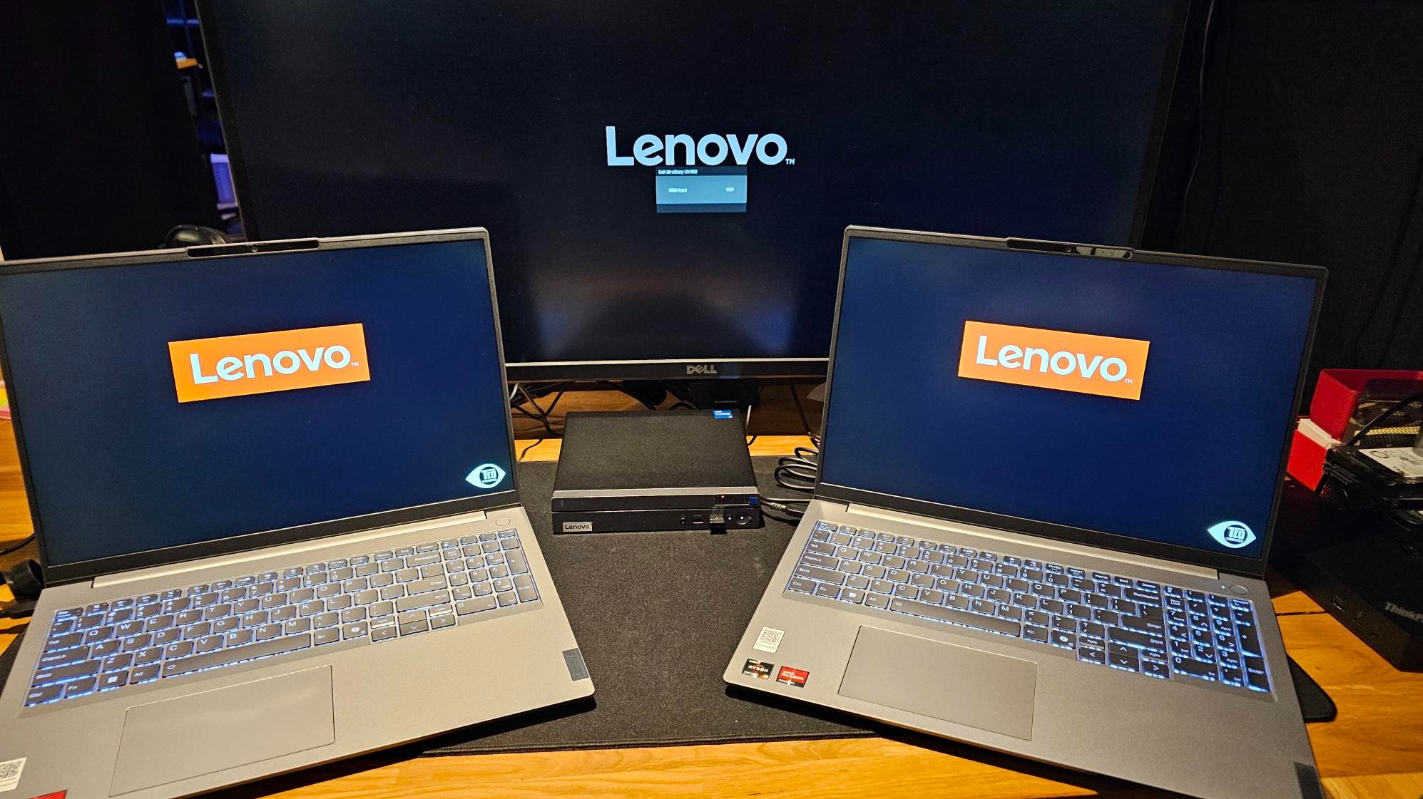 Dwa laptopy Lenovo z podświetlaną klawiaturą, ustawione symetrycznie po bokach monitora Dell, na którym widoczne logo Lenovo. Na środku mały komputer Lenovo.