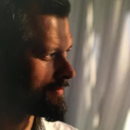 Portret mężczyzny z brodą, profil, ciepłe oświetlenie, w tle okno z zasłonami. Ujęcie z bliska, skupione na wyrazie twarzy.