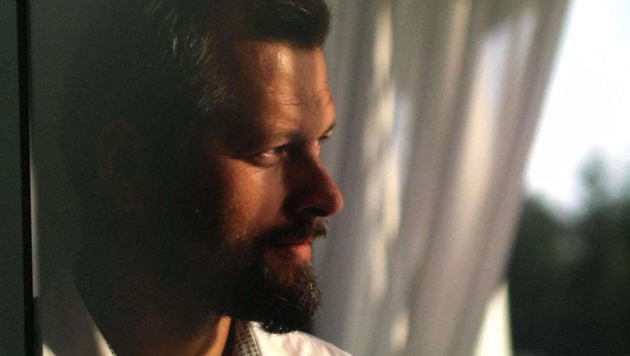 Portret mężczyzny z brodą, profil, ciepłe oświetlenie, w tle okno z zasłonami. Ujęcie z bliska, skupione na wyrazie twarzy.