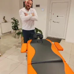 Mężczyzna w białej koszuli stoi obok pomarańczowo-szarego stołu do masażu w jasnym pomieszczeniu z roślinami i drzwiami z napisem Olaplex #myidentity.