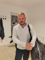 Mężczyzna z krótką brodą i wąsami, ubrany w białą bluzkę z długim rękawem, trzymający na ramieniu dużą, ciemną torbę, prawdopodobnie na sprzęt do masażu, w jasnym wnętrzu z białymi ścianami...