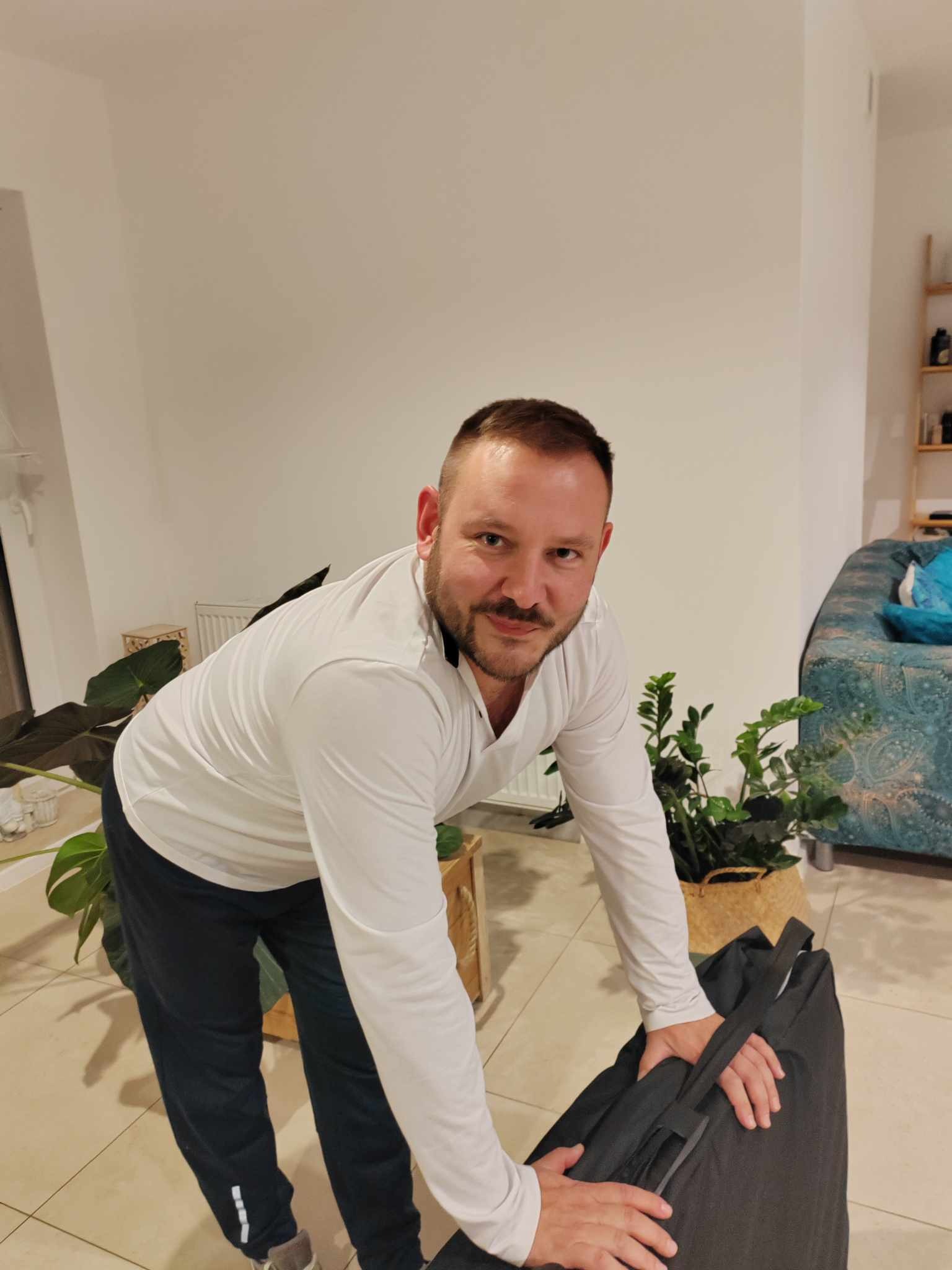 Mężczyzna w białej koszuli i dresach rozpakowuje przenośny stół do masażu w jasnym pomieszczeniu z roślinami doniczkowymi.