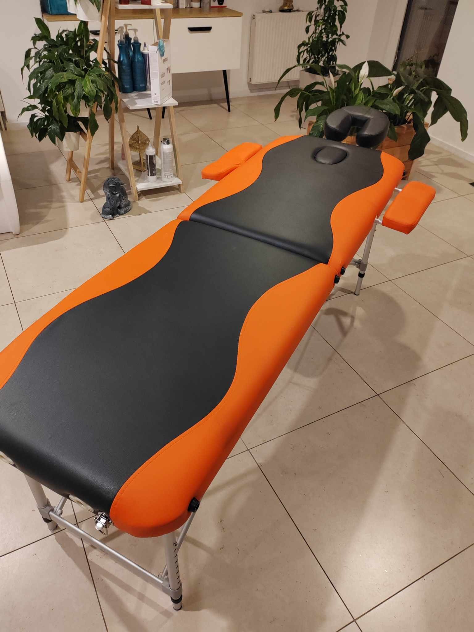 Pomarańczowo-czarne łóżko do masażu z regulowanym zagłówkiem i podłokietnikami, na tle jasnych płytek podłogowych, z roślinami doniczkowymi i meblami w tle.